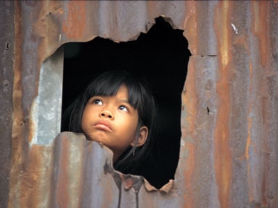 Small child looking through a hole in a tin wall.