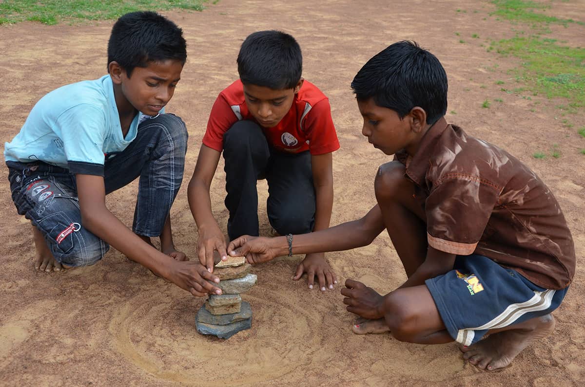 Three boys kneel on the ground, stacking seven stones