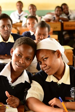 young people in a classroom with two smiling girls touching heads in the foreground