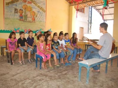 Man sitting on a bench playing guitar for a group of children in a classroom