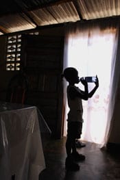 young boy standing in a dark room in front of a window with light streaming in drinking a bottle of water