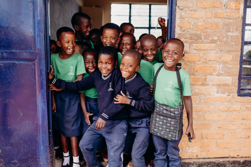 A group of of children wearing school uniforms stand in a doorway and smile and laugh while looking at the camera.