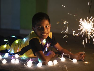 A boy lays on the ground, holding a sparkler and surrounded by Christmas lights.