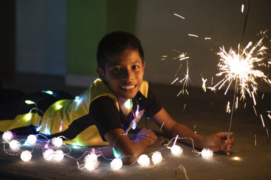 A boy lays on the ground, holding a sparkler and surrounded by Christmas lights.