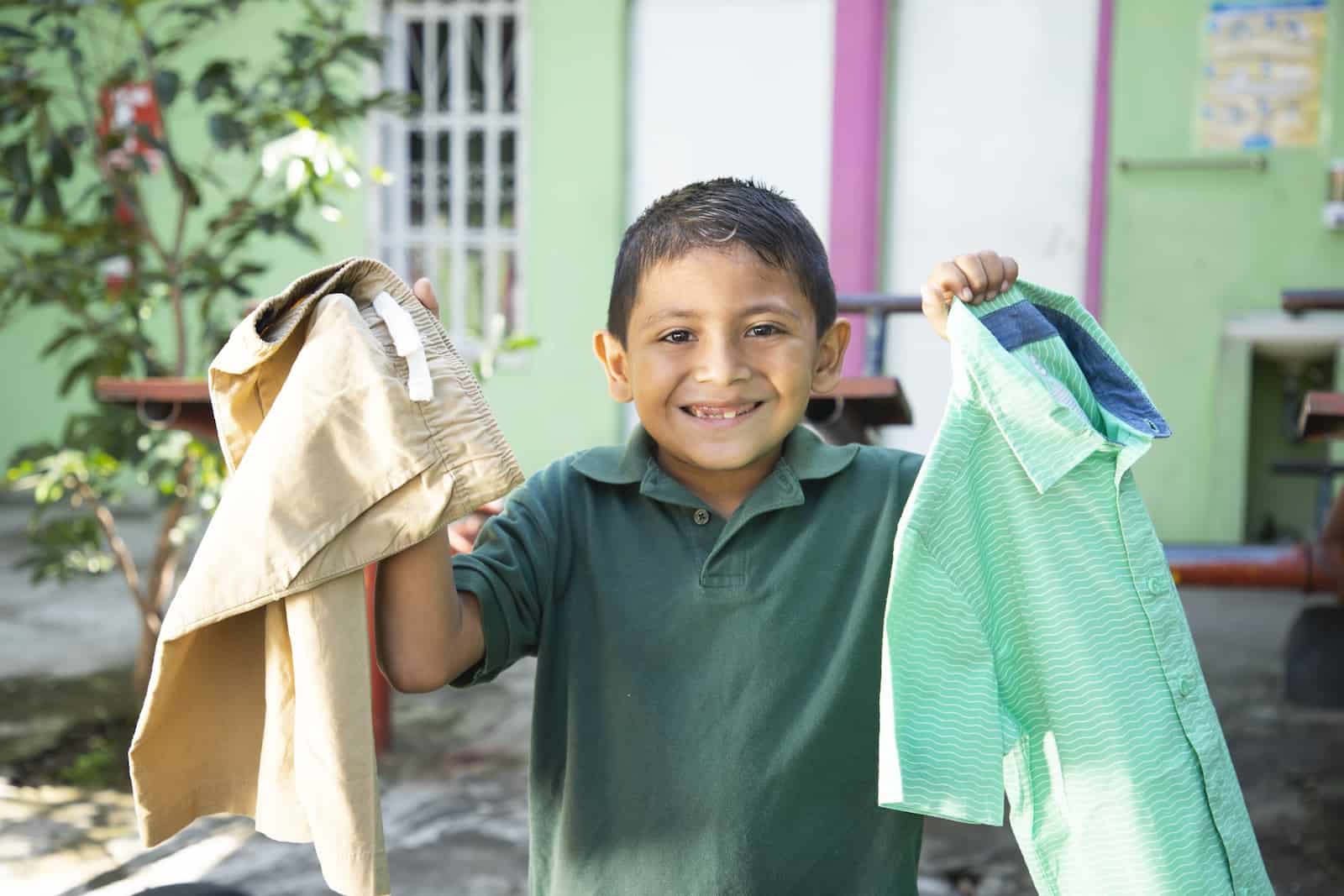 A boy holds up a shirt and pants, smiling. 