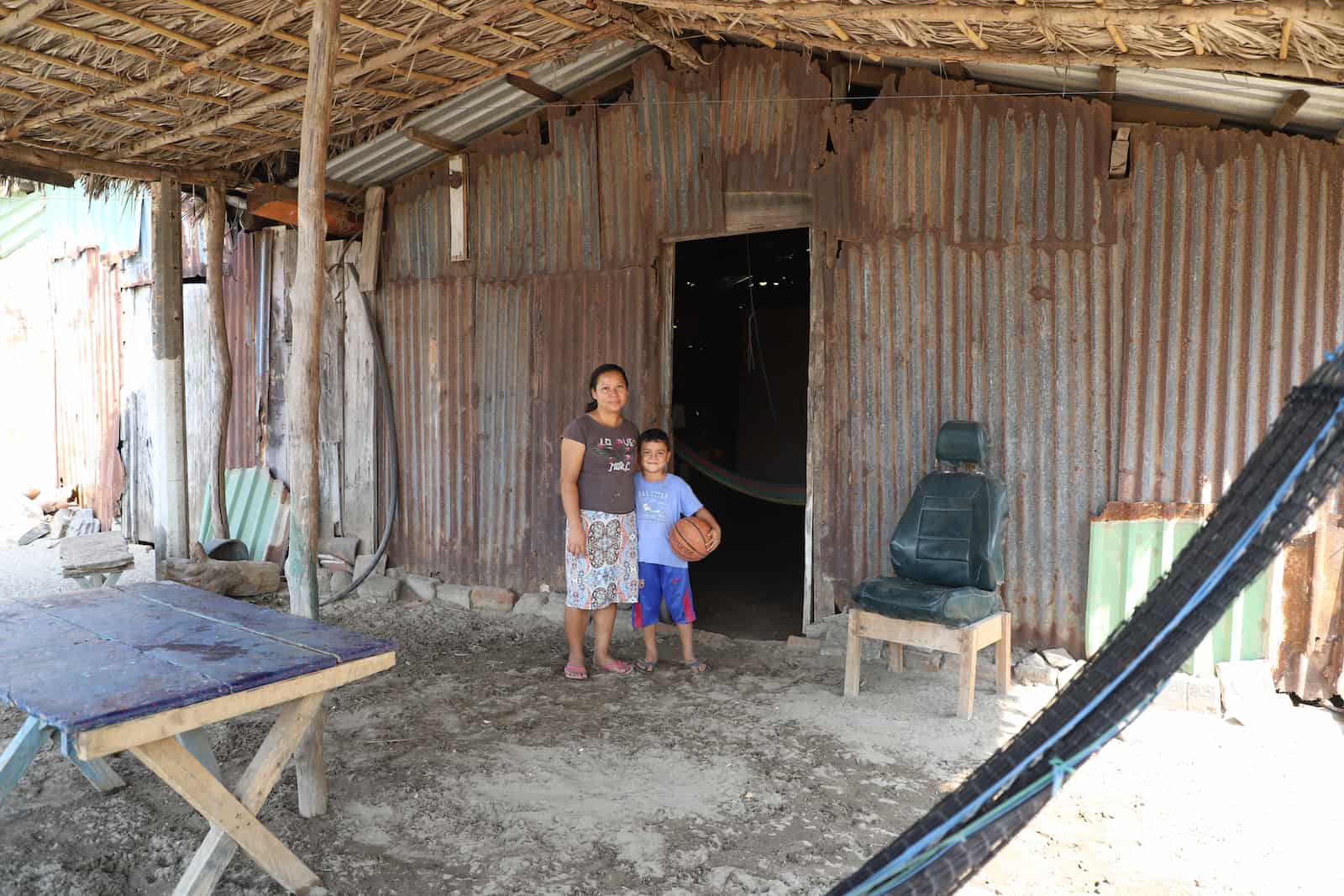 A mom and boy stand in front of a home made from corrugated metal sheets in rural El Salvador