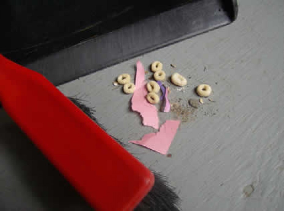 A broom head next to some scraps of pink paper and cheerios on the floor