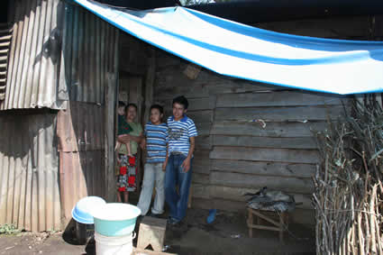 Mother and her three children standing under an awning outside their home made of wood and sheet metal