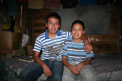 Two brothers sitting on a bed in their home with older brother has his arm around his younger brother