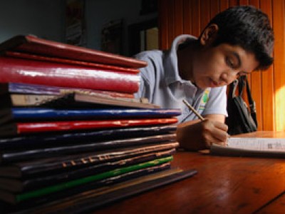 boy sitting and writing beside a stack of books