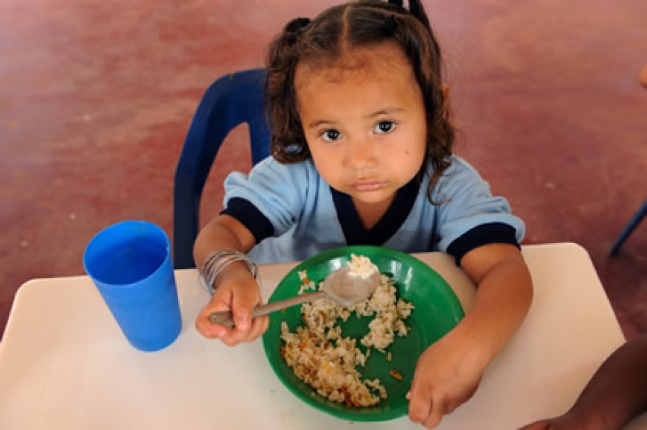 little girl sitting at a table eating rice and beans from a green plate with a blue cup beside her
