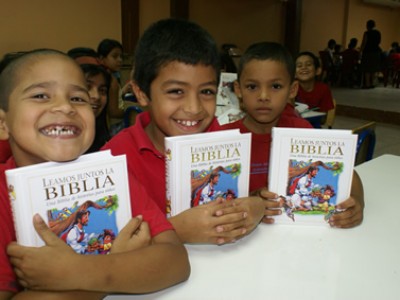 three smiling boys in red shirts holding a bible with their arms resting on a table