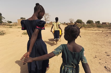 two girls on a dirt road holding hands with another girl in front pushing a wheelbarrow