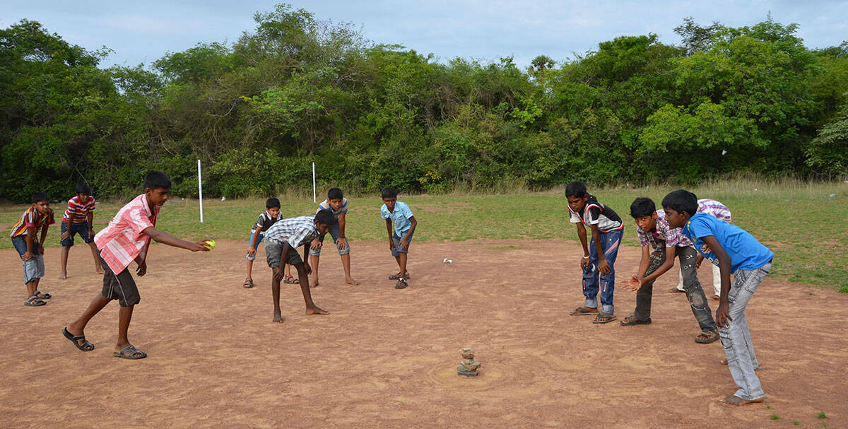 A group of boys stands around a stack of seven stones, one boy throwing a ball towards the stones.