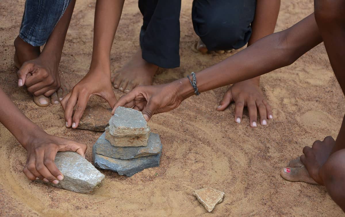 Children crouch on the ground, stacking seven stones