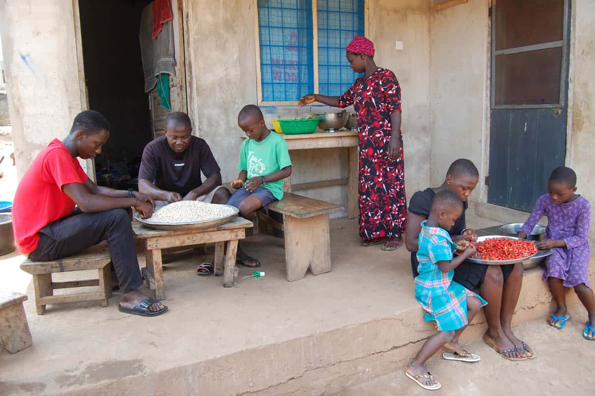 Ghanaians preparing food outdoors. Three children sit on the right preparing red food, while three teens prepare food on the left. A Ghanaian woman is also in the background preparing food.