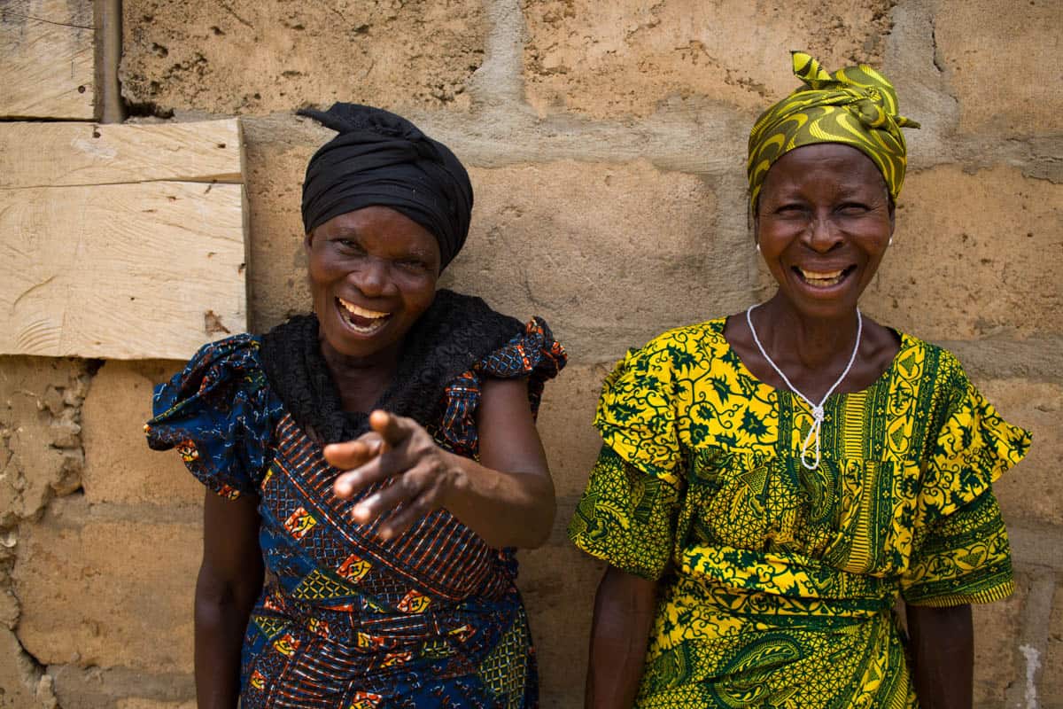 Two women in traditional Ghanaian clothing are sitting next to each other. Both are wearing traditional head wraps. One is laughing and pointing.