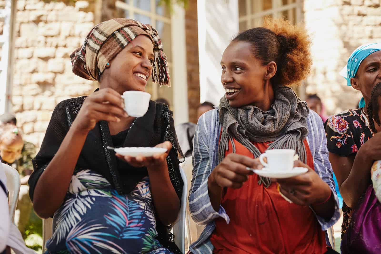 Two women drink coffee, smiling at each other.