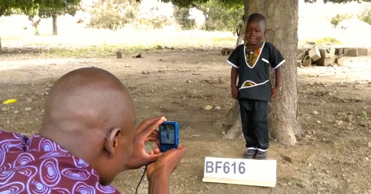 Man taking a picture of a child standing in front of a tree with a sign in front of his feet