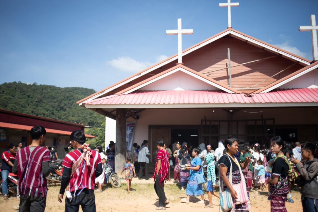 Many people gather outside of a church that features three crosses on the roof.