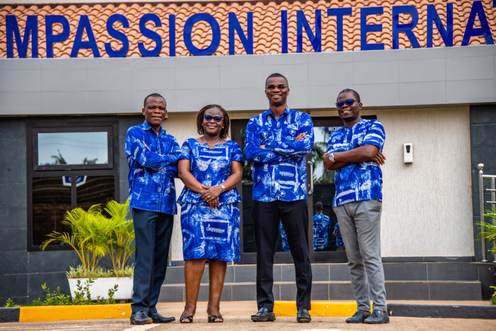 Four people wearing bright blue tops stand in front of a building and smile for the camera.