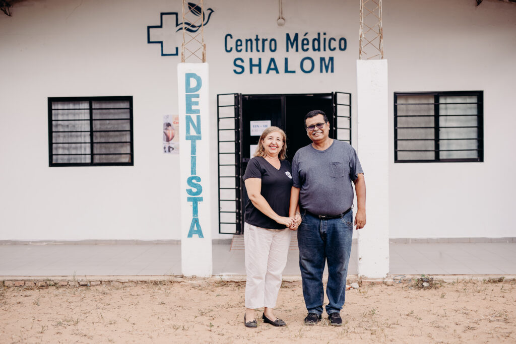 An older man and woman stand in front of a medical clinic while smiling for the camera.