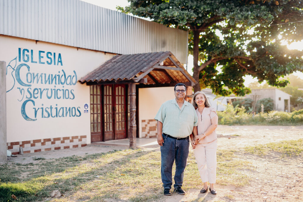 An older man and woman stand outside of a building and smile while the sun shines in from the left.