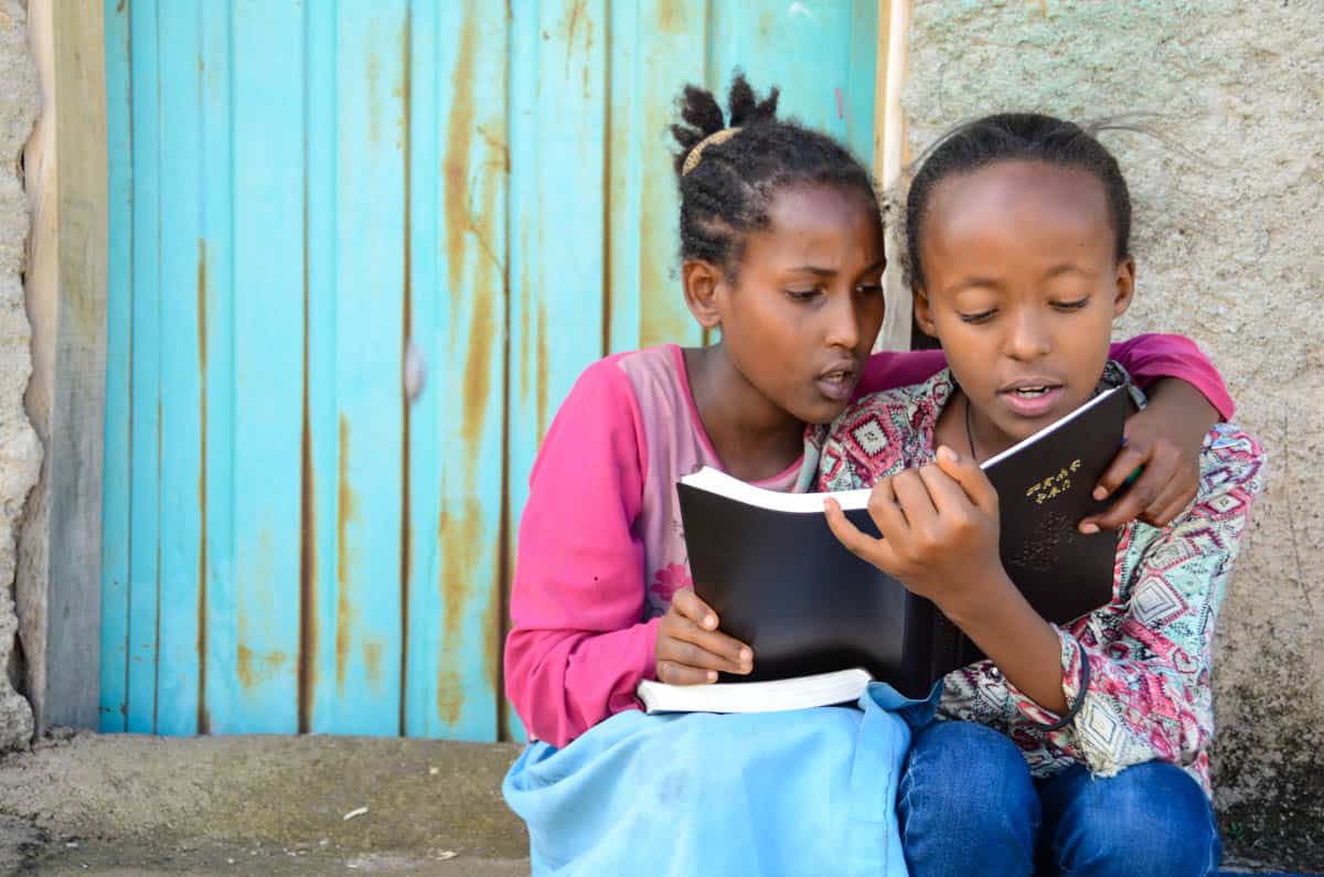 Two girls wearing pink tops and denim bottoms huddle together to read the Bible. 
