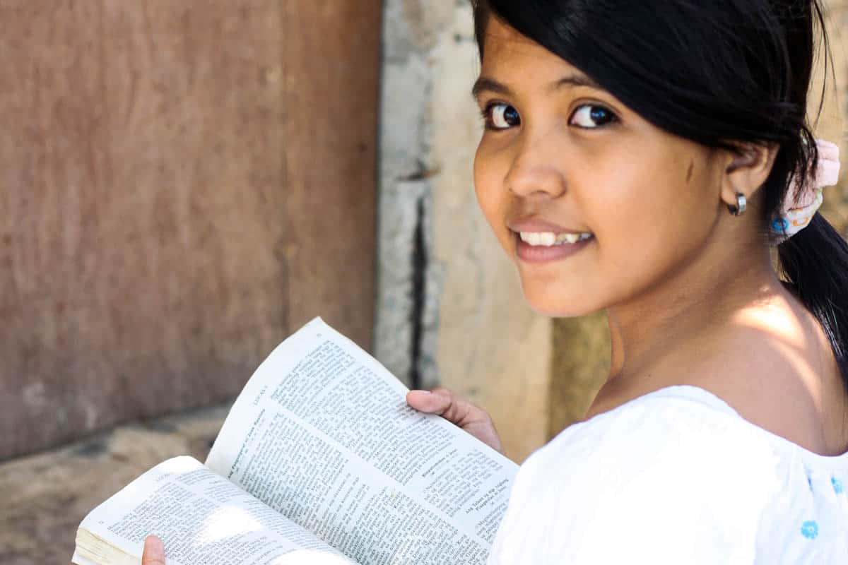 a girl holding an open Bible smiles at the camera. Her dark hair is in a ponytail, and she is wearing a white shirt.