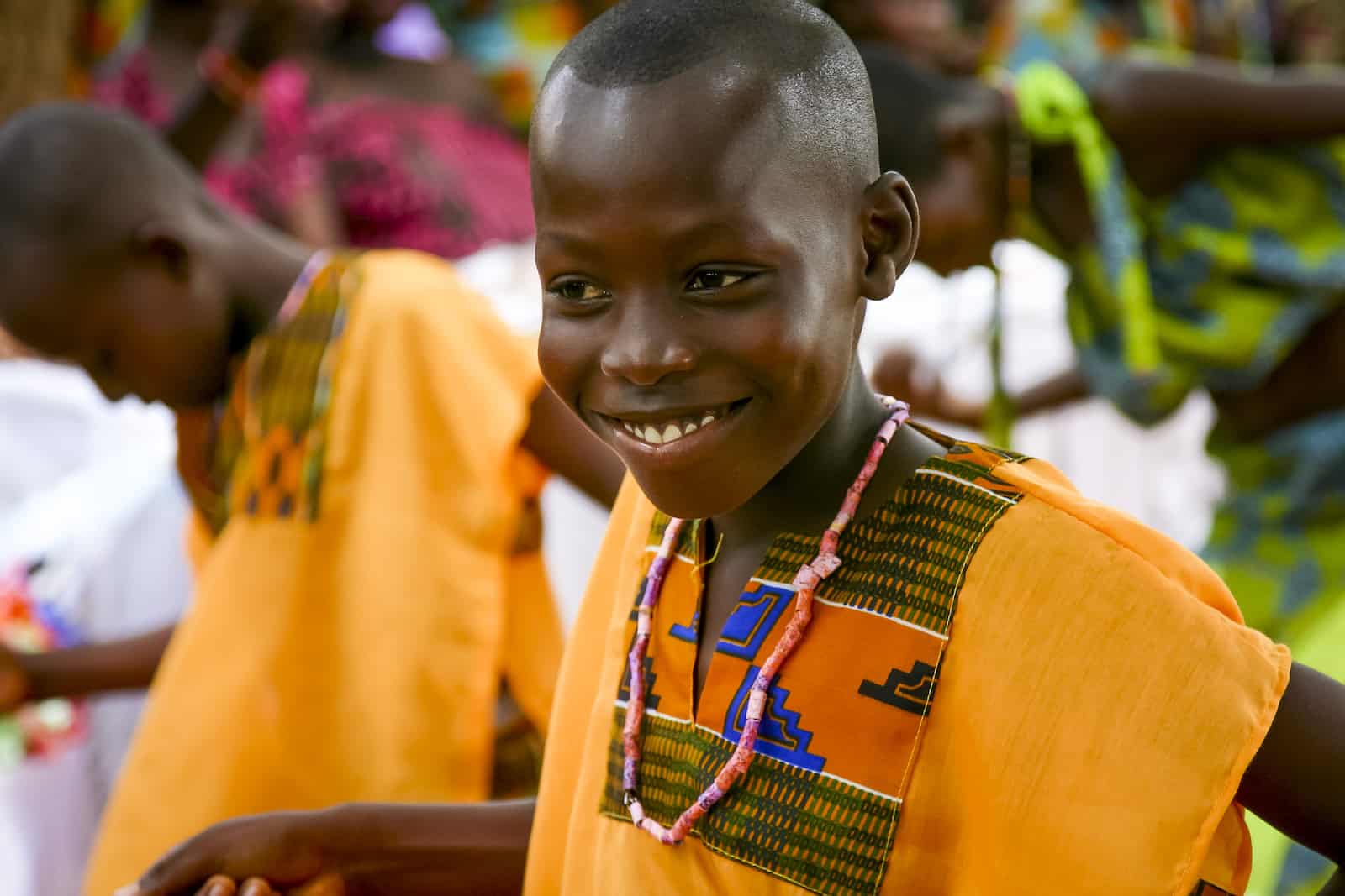 A boy wearing a bright orange and patterned shirt smiles while dancing. Other children can be seen in the background.