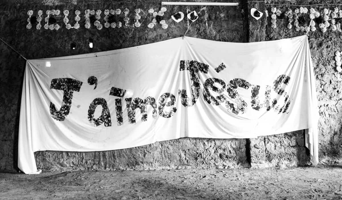 A giant cloth banner, sign reads j'aime Jésus, French for I love Jesus. It is hanging on a mud wall.