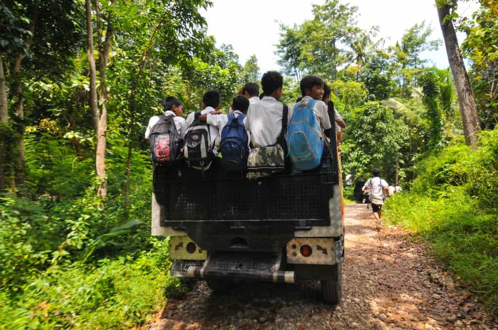 Schoolchildren wearing backpacks ride on the back of a truck down a dirt road in a rural area