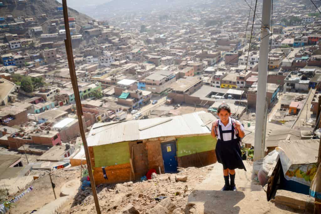 A girl wearing a school uniform and backpack walks up steep stairs. A city is seen in the background and beneath her
