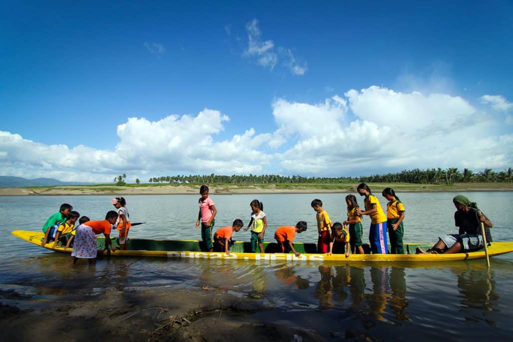 Children ride on a canoe made from a hollowed-out coconut tree. Some are standing and some are sitting. 