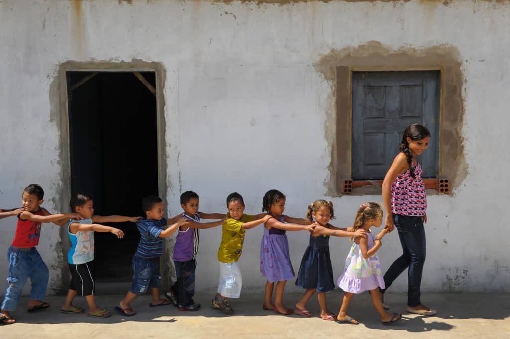 Children line up with their hands on each other's shoulders in single file, following an adult