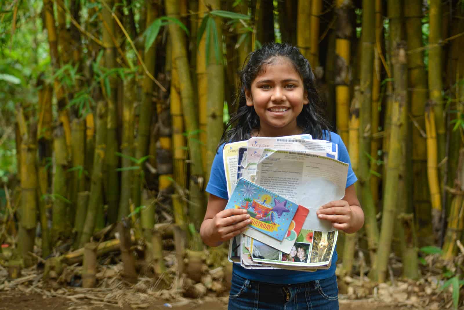 A young girl in a blue shirt and jeans stands in front of a stand of bamboo, holding letters