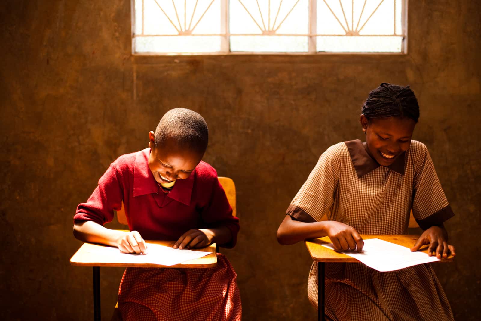 Two girls in brown and red school uniforms sit inside at desks, writing letters