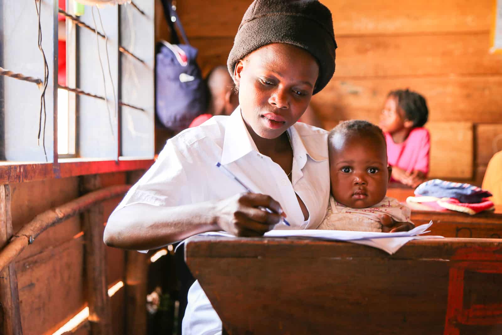 A woman in a white shirt and black hat sits at a desk writing a letter, holding her young child.