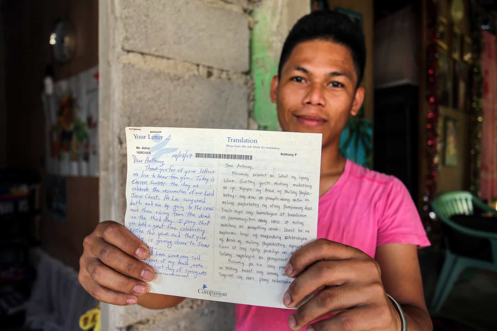 A young man wearing a pink shirt holds up a letter, standing in front of a cement building