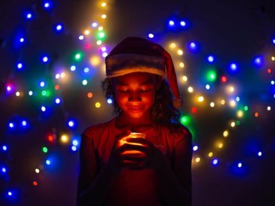 A girl in Brazil wears a Santa hat. She is smiling down at a candle. The room is dimly lit, and Christmas lights sparkle behind her.