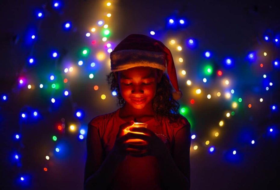 A girl in Brazil wears a Santa hat. She is smiling down at a candle. The room is dimly lit, and Christmas lights sparkle behind her.