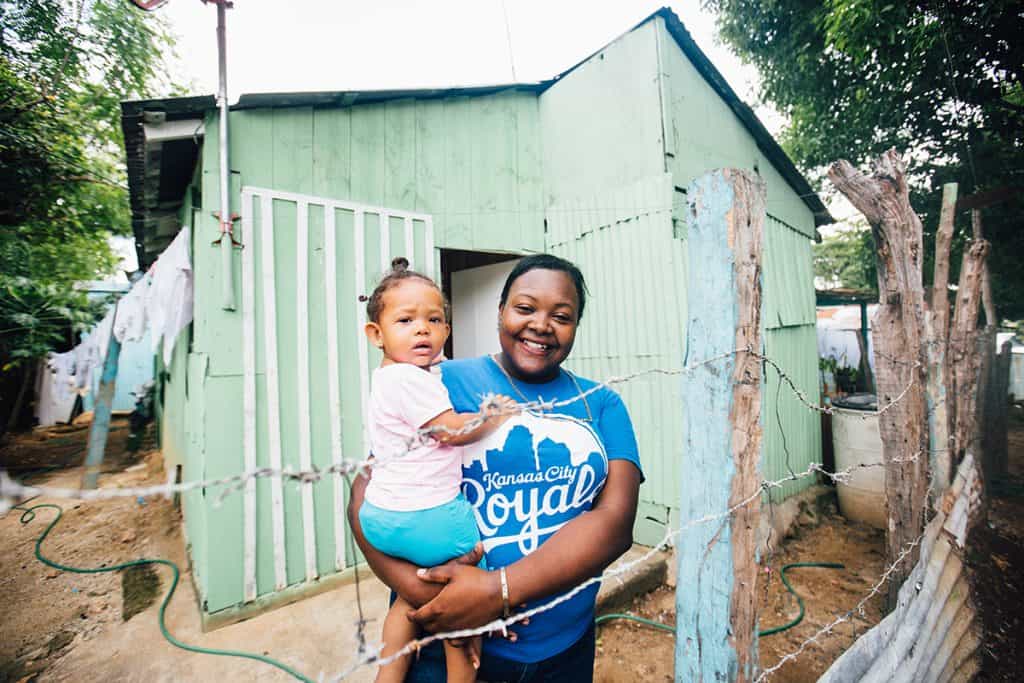 A smiling mother wearing a Royals T-shirt holds a small girl child. They are standing outside a pastel green structure where they live