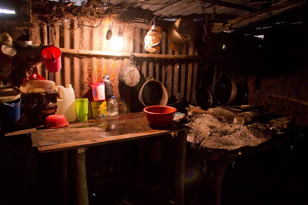 In this tiny kitchen, pots and pans hang from the roof and tin walls of a tiny home. A small wooden table is seen next to a pile of ashes where the family cooks.