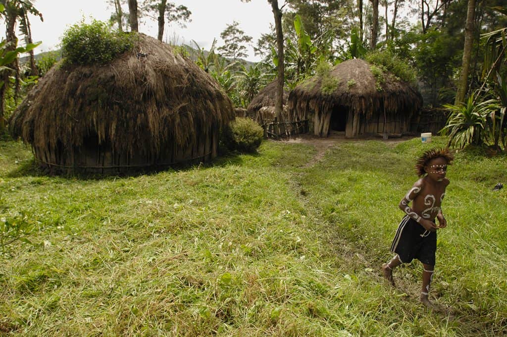 A boy in Indonesia running in the grass outside his tiny home, made of wooden slats and thatched roof.