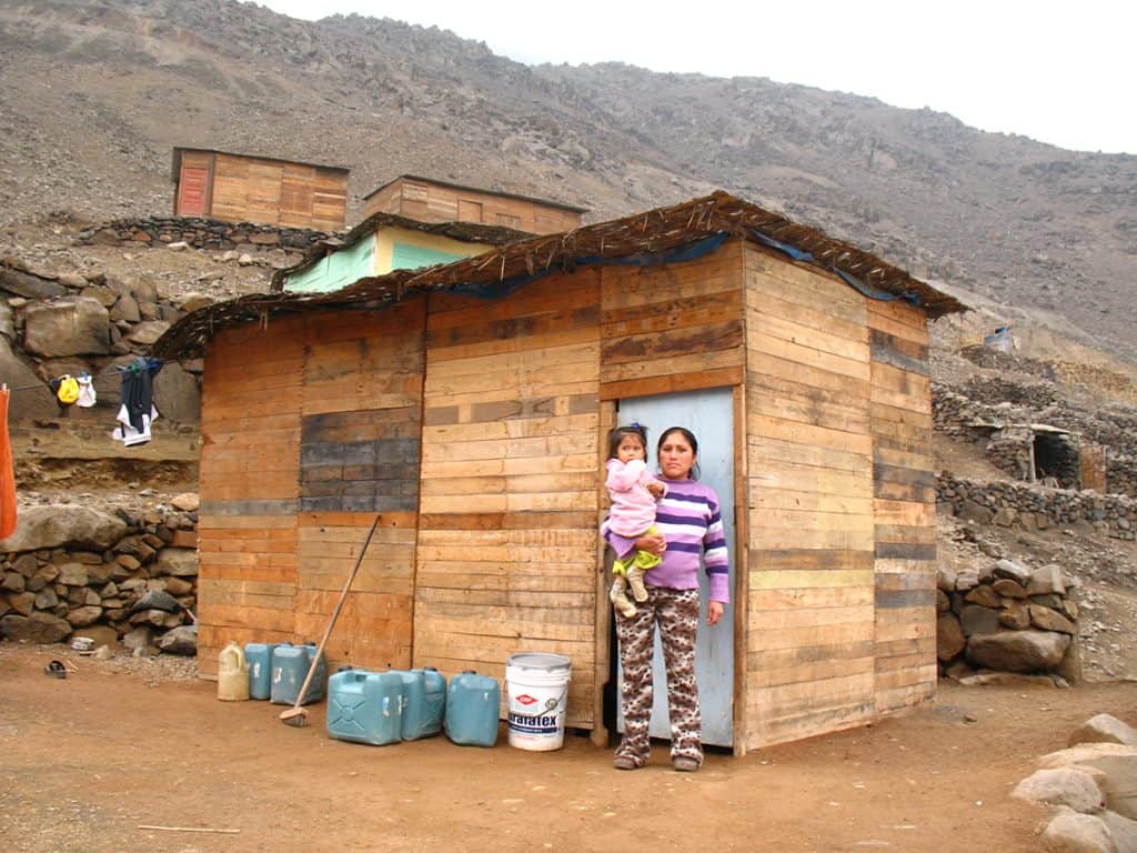 A mother holds a young girl. They are standing outside a small wooden home in Peru. Mountains are seen in the background.