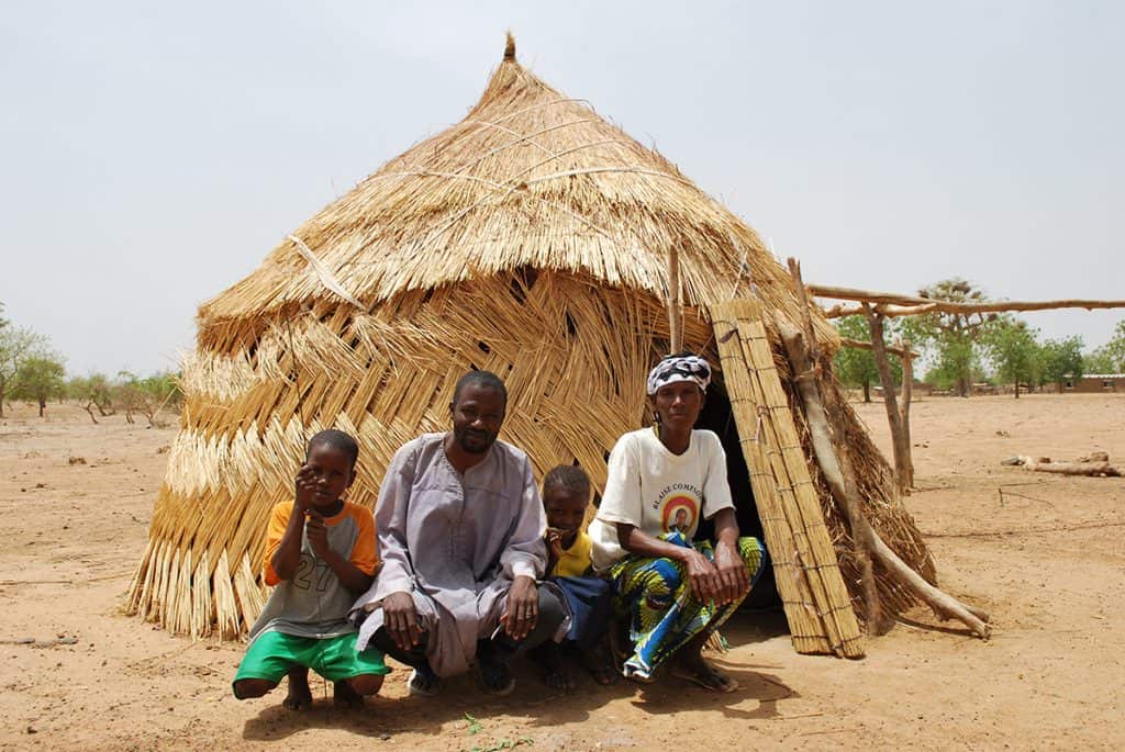 A family, a father and mother with two children, sit outside their home in Burkina Faso, which is made of woven reeds. It is light tan in color.