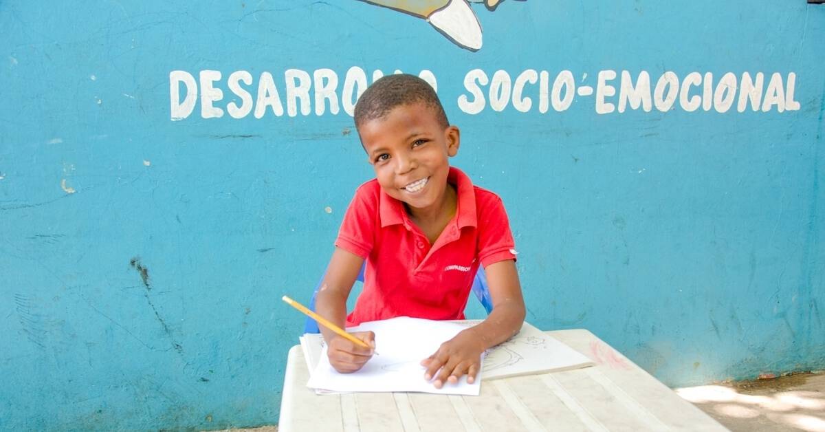 smiling boy in red shirt sitting at desk writing a letter