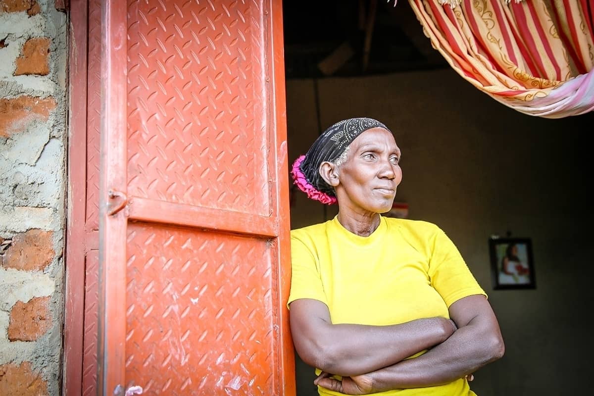 A woman, Grace's mother, stands in the doorway of their home in Uganda wearing a yellow shirt