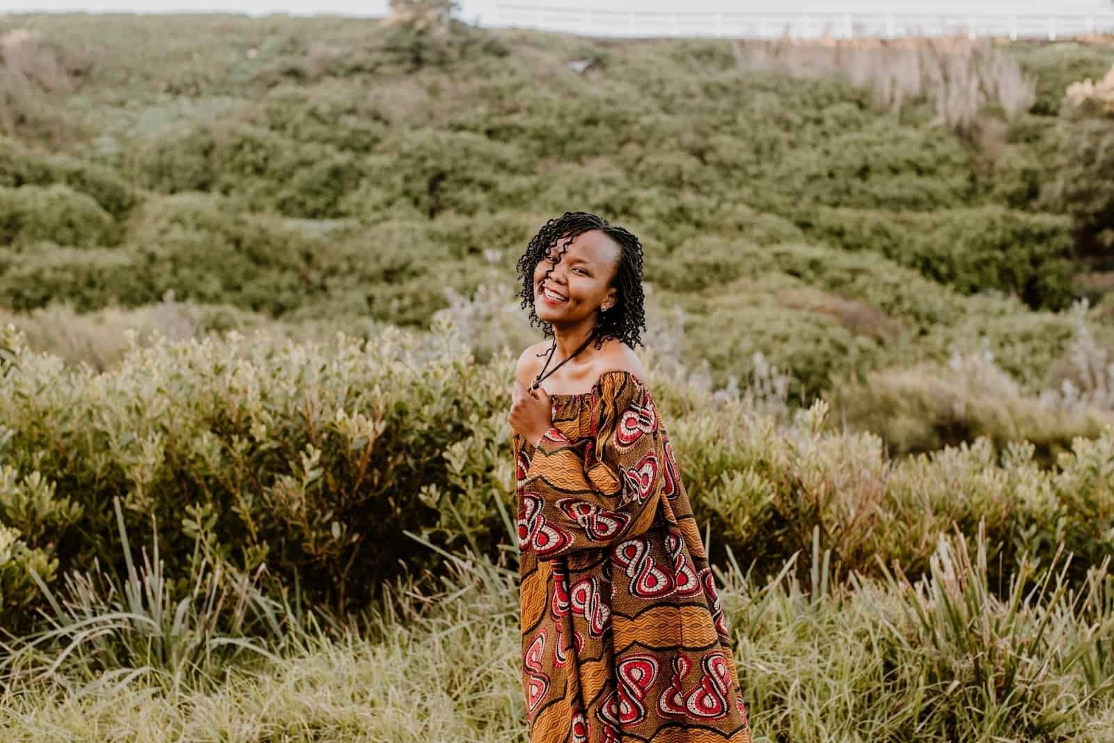 A woman, Christine, stands in a green field, smiling, wearing a traditional African dress and holding the necklace she wears around her neck.