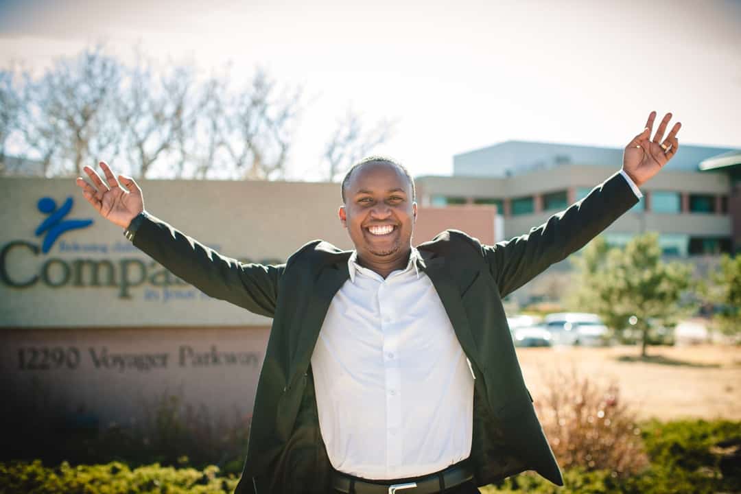 Jean Claude stands with his arms out to the side with a large smile on his face in front of a building with a sign that says Compassion International.